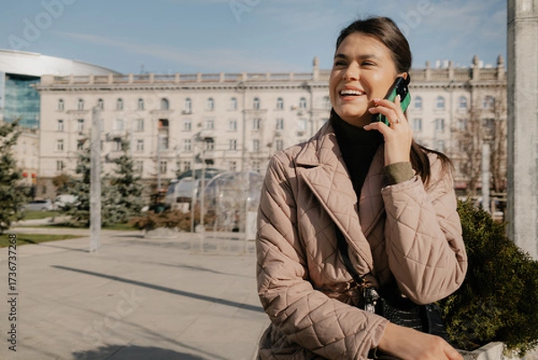 Fototapeta Cheerful woman chats on her mobile outdoors in an urban setting with modern architecture and blue sky.