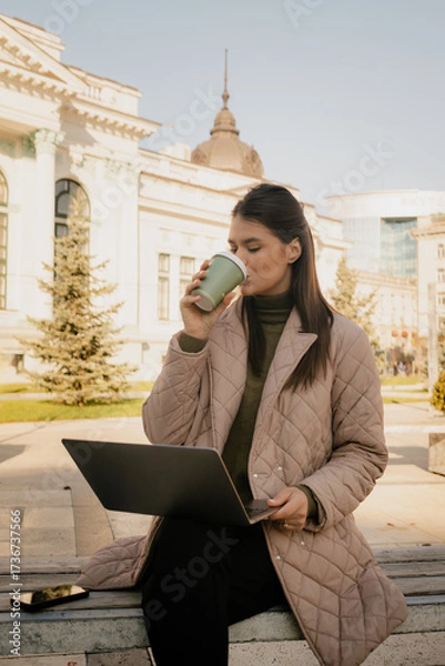 Fototapeta Professional woman uses laptop outside, enjoying coffee in an urban plaza with historic architecture.