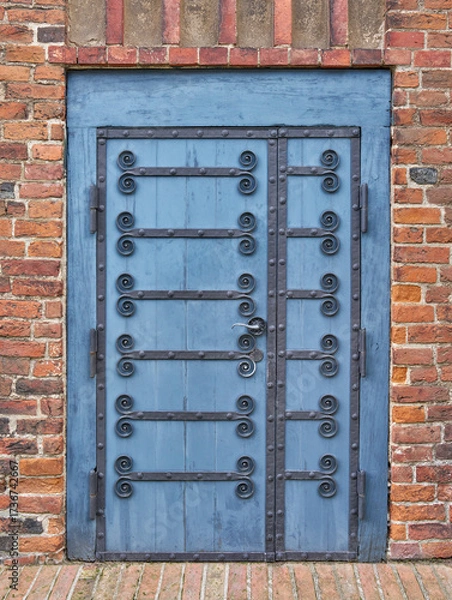 Obraz Blaue Tür mit Rahmen und kunstvollen Verzierungen aus Metall in einer roten Backsteinfassade