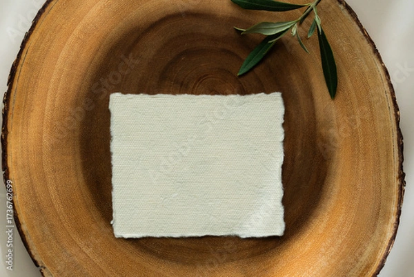 Fototapeta Blank textured paper card on wooden slice with olive branch on beige background. Minimalist mockup with natural light and shadows.	