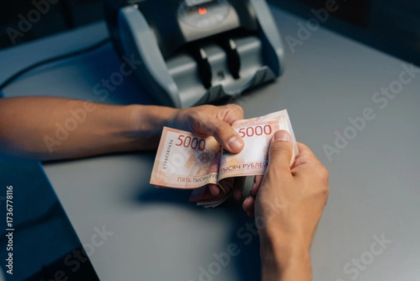 Fototapeta Close-up cropped shot of cashier male counting Russian rubles banknotes late at night after closing store using money counting machine in background. Concept of financial accounting in modern office.