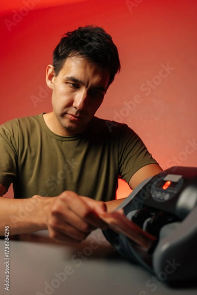 Fototapeta Vertical studio portrait of serious focused cashier counting stack of banknotes using professional electronic money counting machine with red light on red isolated background.