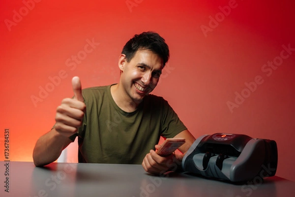 Fototapeta Successful cashier giving thumbs up while counting banknotes with money counter machine sitting at gay table, smiling looking at camera, radiating positivity and success, on isolated red background.
