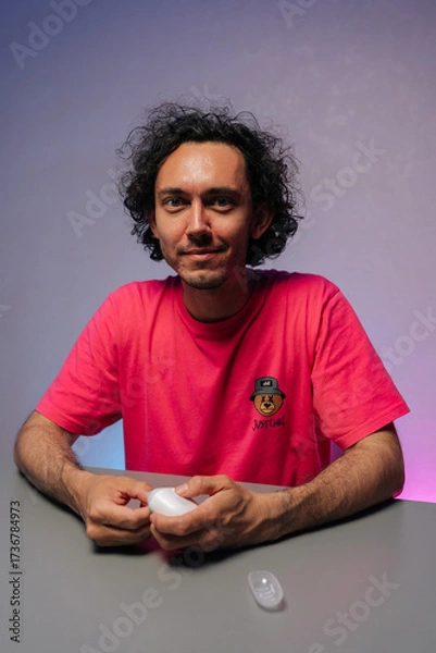 Fototapeta Vertical portrait of handsome young adult man with curly hair and pink t-shirt cutting fingernails using nail clipper sitting at gray table against purple background, smiling looking at camera.
