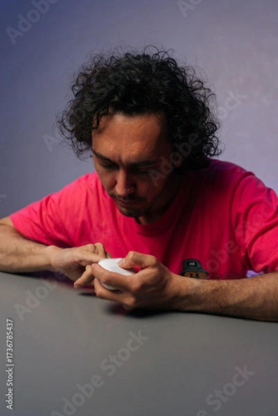 Fototapeta Vertical portrait of focused young adult male with curly hair trimming fingernails using nail clipper, sitting at gray table with purple backdrop at home. Concept of healthy lifestyle.