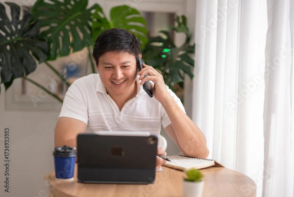 Fototapeta Asian freelancer talking on phone and taking notes while working on tablet at home