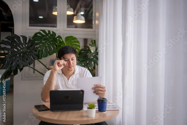 Fototapeta Asian freelancer working from coffee shop reading documents and using tablet