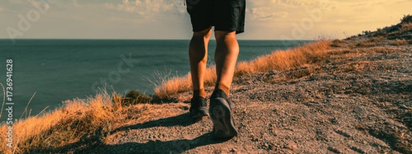 Fototapeta A man takes a brisk walk outdoors on a trail located on an elevated area overlooking the sea or ocean. A close-up view from behind.