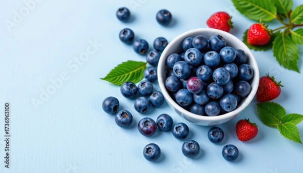 Fototapeta Fresh blueberries in white bowl surrounded by vibrant strawberries and green leaves create delightful and colorful display. This arrangement evokes sense of freshness and healthiness