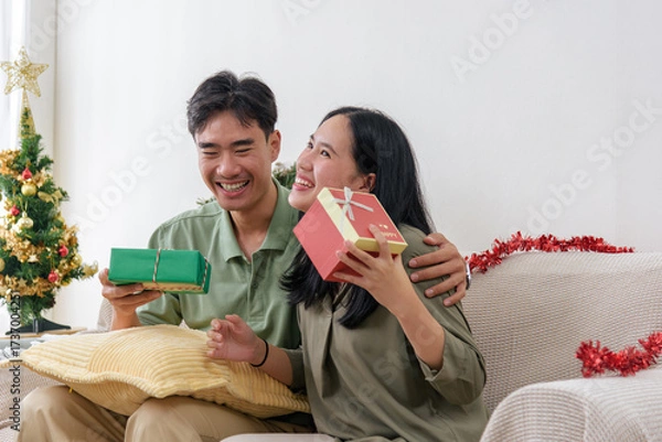 Fototapeta Cheerful pair exchanging wrapped presents during christmas holidays, smiling as they sit near an ornamented fir tree, holiday joy and festive celebration are evident
