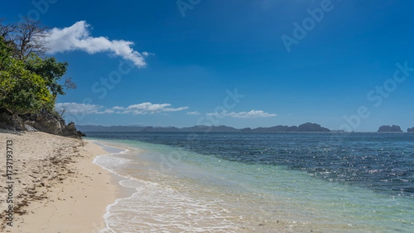 Fototapeta A beautiful secluded tropical beach with no people. A calm turquoise ocean. Foam of waves on white sand. Green vegetation, boulders on the shore. Mountains on the horizon. Clear blue sky. Copy space. 