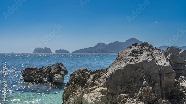 Fototapeta Picturesque cliffs in the ocean. Reflections on the surface of the turquoise sea. Mountains on the horizon. In the foreground is a rock with rough furrowed slopes. Clear blue sky. Copy space.