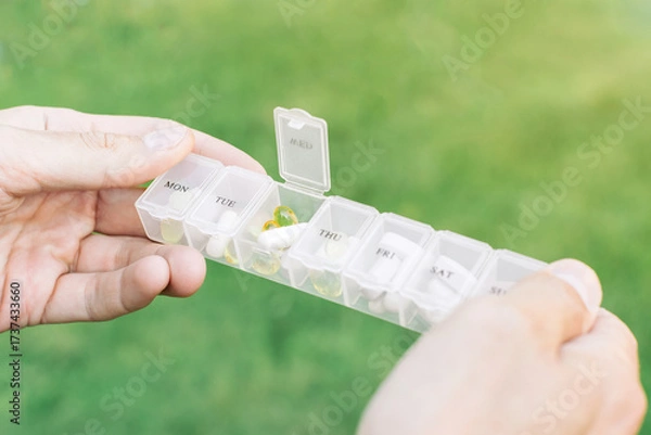 Fototapeta Weekly pill organizer with compartments for each day of the week filled with capsules and tablets held in hands against green outdoor background
