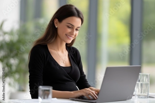 Fototapeta Pregnant woman in black top smiles while working on laptop at bright office desk with glass of water, showcasing a modern workspace atmosphere and professional engagement