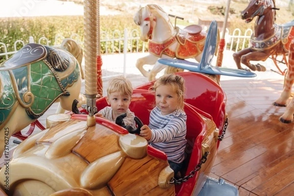 Obraz A girl and a boy ride on a carousel, in a car. Happy kids