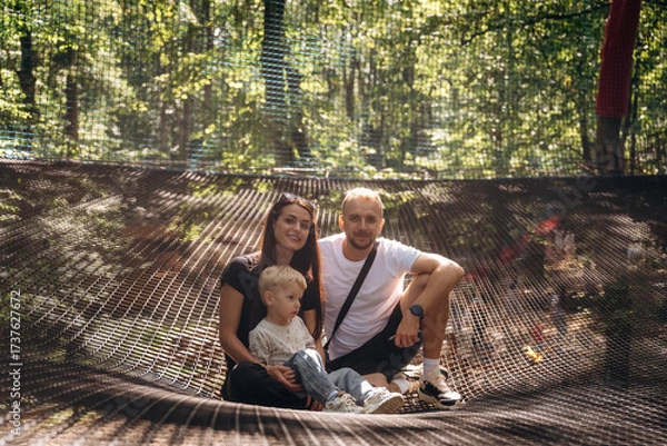 Obraz Family of father, mother and little son sitting on the children's amusement ropes course