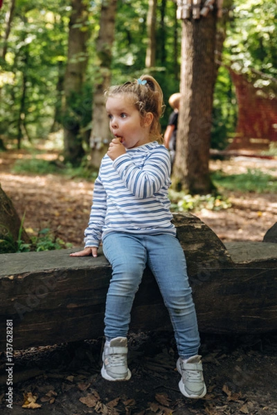 Obraz Funny little girl is sitting on the wooden bench and eating ice cream. In the public park