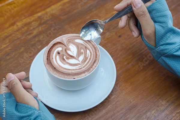 Fototapeta Cappuccino coffee with heart-shaped latte art on a wooden table, a close-up photo of a cappuccino drink in a white cup with a spoon on a wood background.