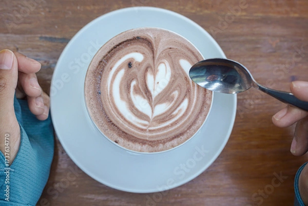 Fototapeta Cappuccino coffee with heart-shaped latte art on a wooden table, a close-up photo of a cappuccino drink in a white cup with a spoon on a wood background.