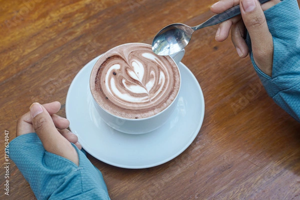 Fototapeta Cappuccino coffee with heart-shaped latte art on a wooden table, a close-up photo of a cappuccino drink in a white cup with a spoon on a wood background.