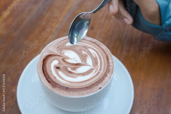 Fototapeta Cappuccino coffee with heart-shaped latte art on a wooden table, a close-up photo of a cappuccino drink in a white cup with a spoon on a wood background.