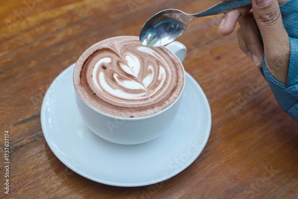 Fototapeta Cappuccino coffee with heart-shaped latte art on a wooden table, a close-up photo of a cappuccino drink in a white cup with a spoon on a wood background.