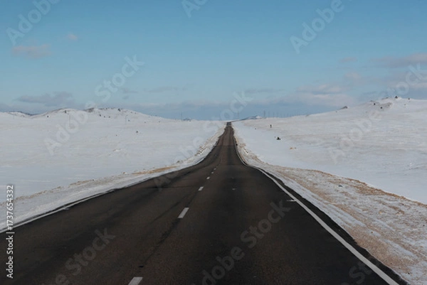 Fototapeta Empty highway stretching into the distance through snow-covered hills under a clear blue sky, symbolizing travel and solitude
