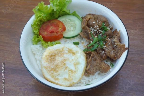 Obraz A vibrant bowl of rice topped with grilled beef, a fried egg, and colorful vegetables, served in a rustic dish