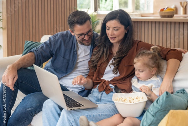 Fototapeta Happy family at home. Mother, father and little daughter on the sofa with a laptop and a bowl of popcorn, relaxing and smiling while enjoying quality time together love, togetherness and simple living