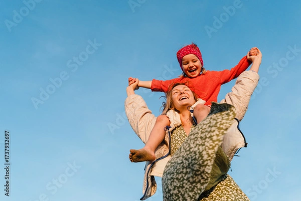Fototapeta Mother carrying her daughter on her shoulder against blue sky. Smiling mom carrying happy little girl making airplane arms. Young family have fun at beach in windy winter.