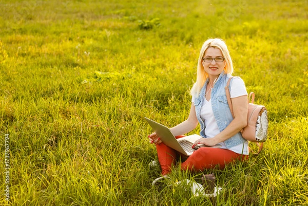 Fototapeta Young woman with laptop sits on the grass in the park on a sunny day. Laughing blonde in a white tank top and shorts. Online training, remote work and communication in social networks