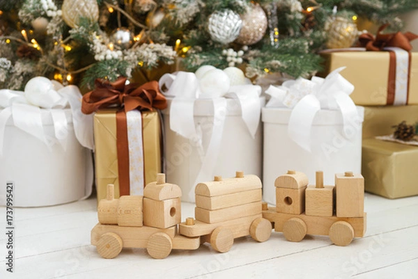 Fototapeta Wooden toy train with wagons placed on white floor in front of wrapped Christmas gift boxes and decorated tree with ornaments and shining lights. Atmospheric mood of winter holidays, New Year