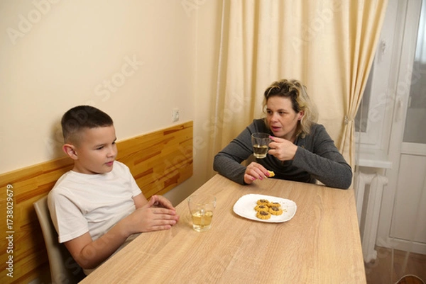 Obraz Family moment with mother and son sitting at table, drinking tea, eating cookies, and enjoying warm conversation together indoors.