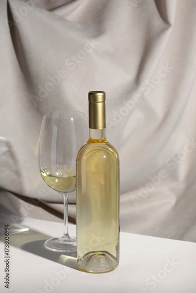 Fototapeta White Wine bottle mockup with glass of white wine on a minimalist table, featuring soft fabric backdrop, showcasing elegant design and inviting atmosphere for branding concepts