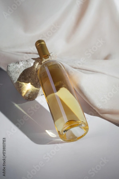 Fototapeta White Wine bottle mockup featuring golden liquid, elegantly positioned on textured stone, with soft fabric backdrop, ideal for branding and design purposes with copy space