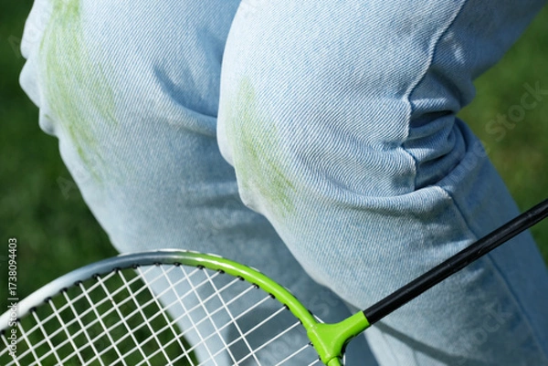 Fototapeta A player in blue jeans kneels on the grass during a break in a game. Green stains visible on the knees of the jeans, showing the playfulness of the outdoor badminton match