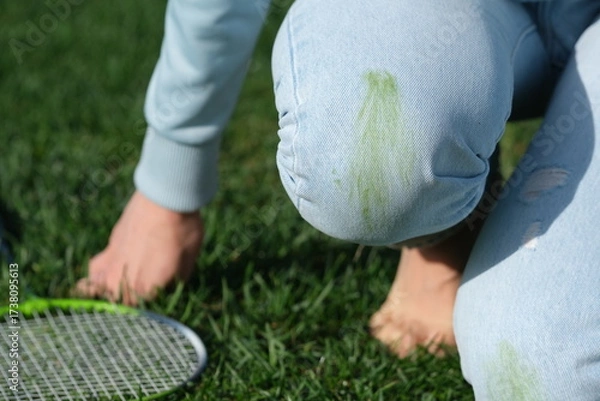 Fototapeta A player in blue jeans kneels on the grass during a break in a game. Green stains visible on the knees of the jeans, showing the playfulness of the outdoor badminton match