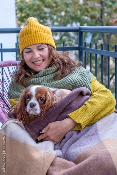 Fototapeta Woman in cozy sweater and hat enjoys autumn on balcony, wrapped in blanket with dog, showcasing warmth, companionship, and comfort during menopause season