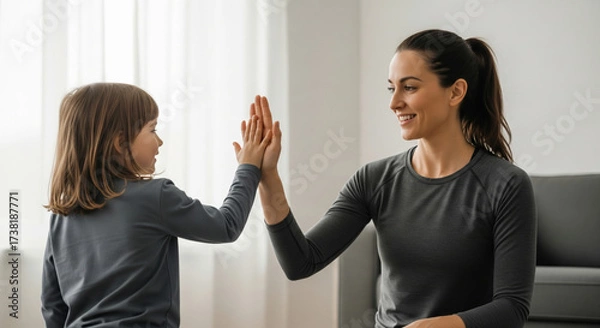Obraz Woman and young girl share high five in bright living room, symbolizing active lifestyle and wellness. Their interaction reflects discipline and positivity in family setting