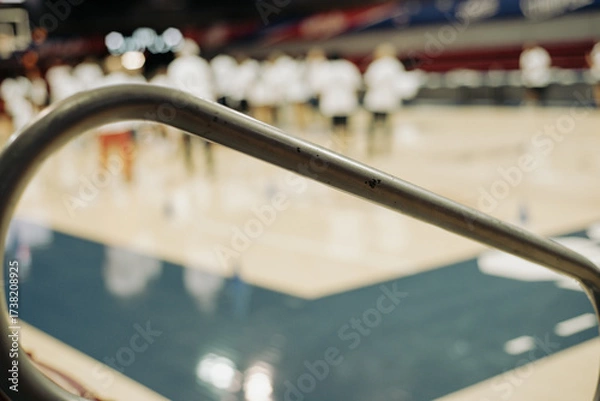 Fototapeta Curved steel handrail in sharp focus leads into a basketball college stadium in Dallas, Texas. Blurred figures in white shirts move across hardwood, blending infrastructure with athletic energy