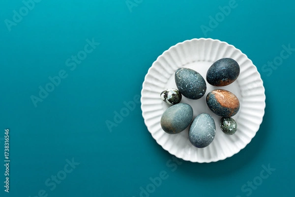 Fototapeta Decorative Easter eggs with various textures and colors are arranged neatly on a white plate, resting on a vibrant teal surface, creating a festive spring atmosphere
