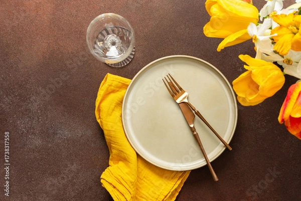Fototapeta A beautifully arranged spring table setting features a light plate, gold-tone utensils, and vibrant flowers. The yellow napkin adds a pop of color, creating a joyful atmosphere for dining