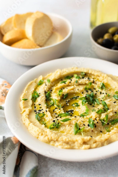 Fototapeta A bowl of creamy hummus topped with olive oil and herbs sits beside fresh bread and a bowl of olives. The light setting showcases a simple, inviting meal