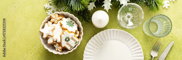 Fototapeta A bright green Christmas table setting with an empty plate and cutlery. The table is decorated with spruse branches and white tree ornaments, creating a festive vibe, banner
