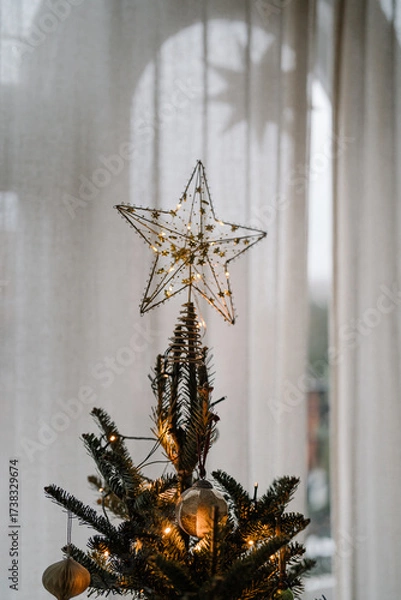 Fototapeta Decorated Christmas tree with a golden star topper and glowing fairy lights against soft curtains.