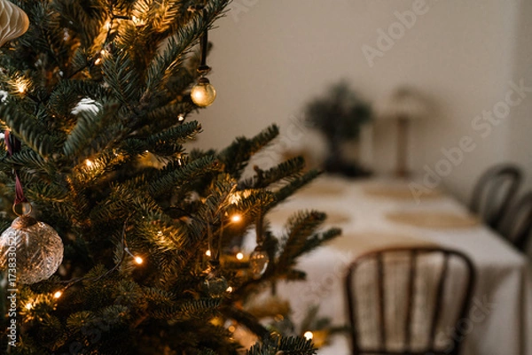 Fototapeta Minimalist Christmas tree with golden ornaments and warm fairy lights in a cozy dining room setting.