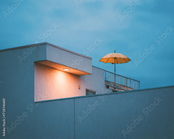 Fototapeta A modern building with a balcony adorned by a bright yellow umbrella, set against a moody blue sky at dusk.