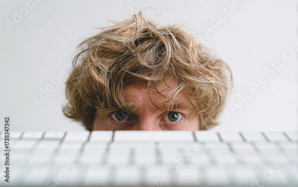 Fototapeta A focused individual with messy hair peers over a keyboard, capturing a moment of concentration in a minimalist workspace.