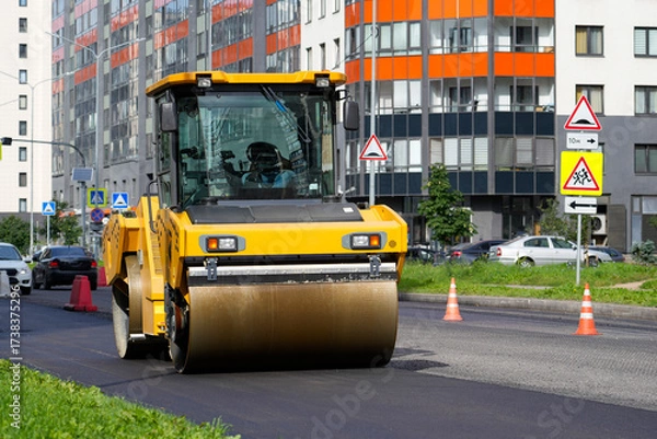 Fototapeta Heavy machinery is engaged in road construction as a steamroller flattens new asphalt while drivers navigate traffic cones in an urban setting with skyscrapers