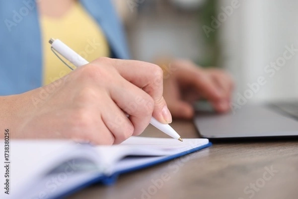 Fototapeta Woman taking notes while passing online test on laptop at wooden table indoors, closeup